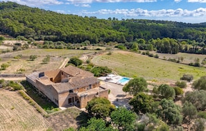 A rustic stone building with a tiled roof, a swimming pool, and a surrounding landscape with trees and open fields.