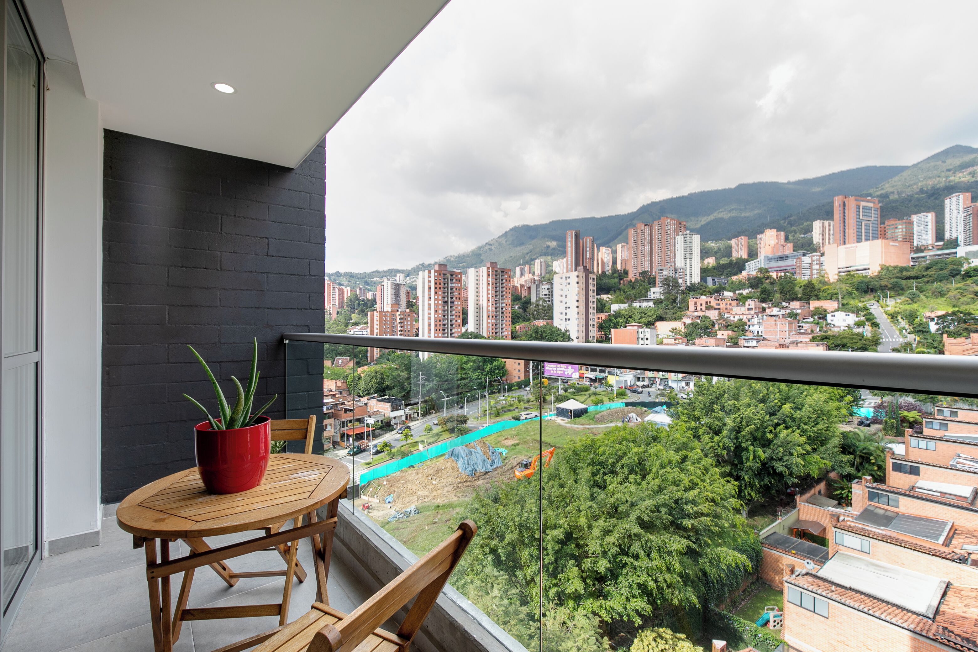 A balcony with a wooden table and chairs, a red pot with a plant, and a view of a cityscape with buildings and mountains.