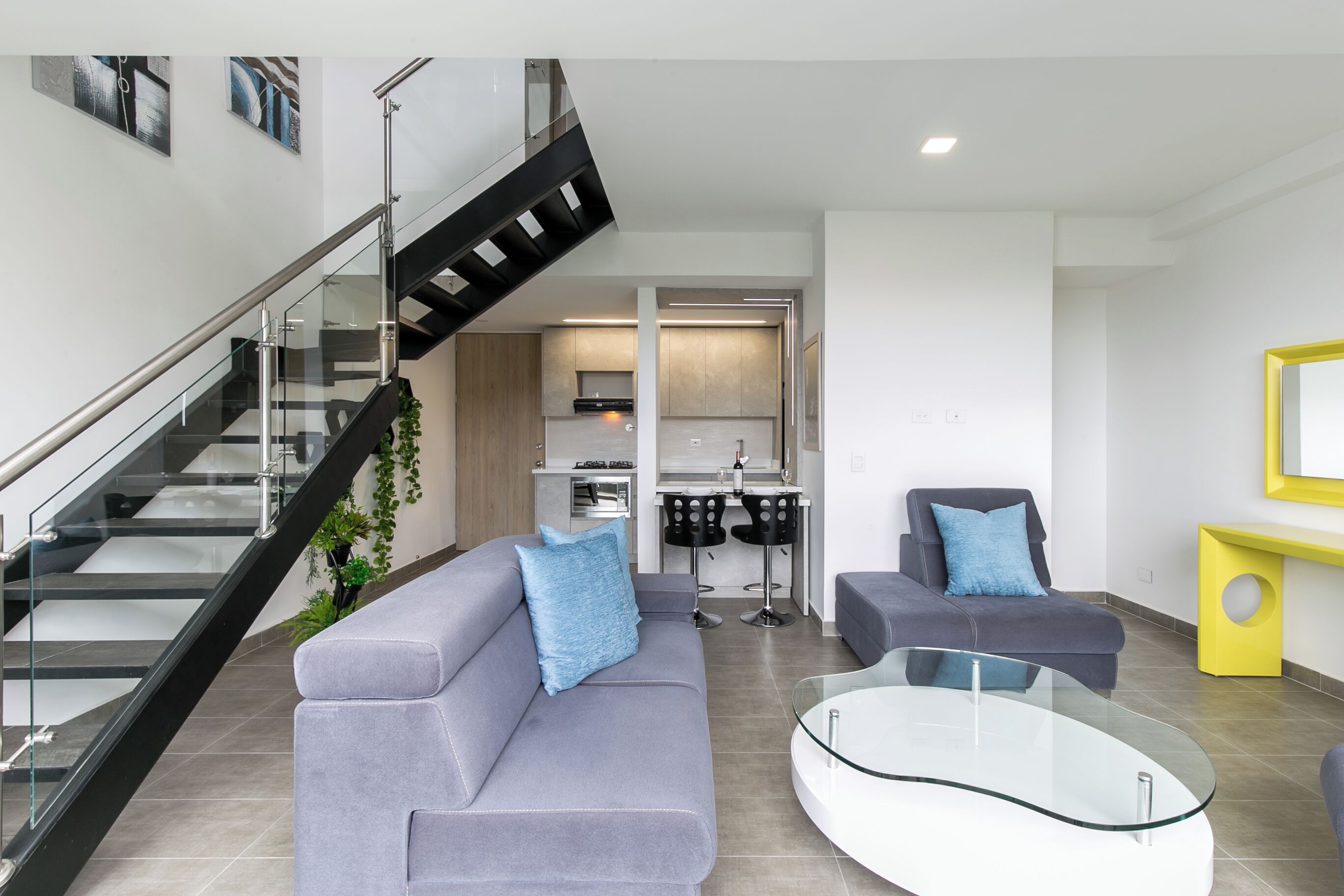 A modern living room with a glass staircase, grey sofas, a glass coffee table, and a kitchen area in the background.