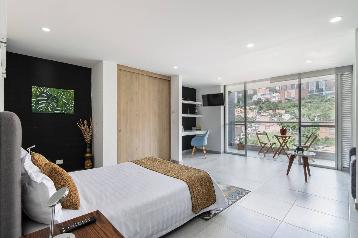 A modern hotel room with a bed, a desk, a chair, a TV, and a balcony with a city view.
