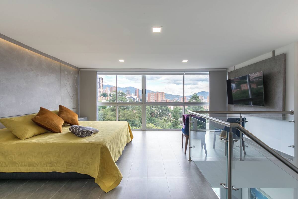 A modern hotel room with a large bed, a balcony with a glass railing, and a city view.
