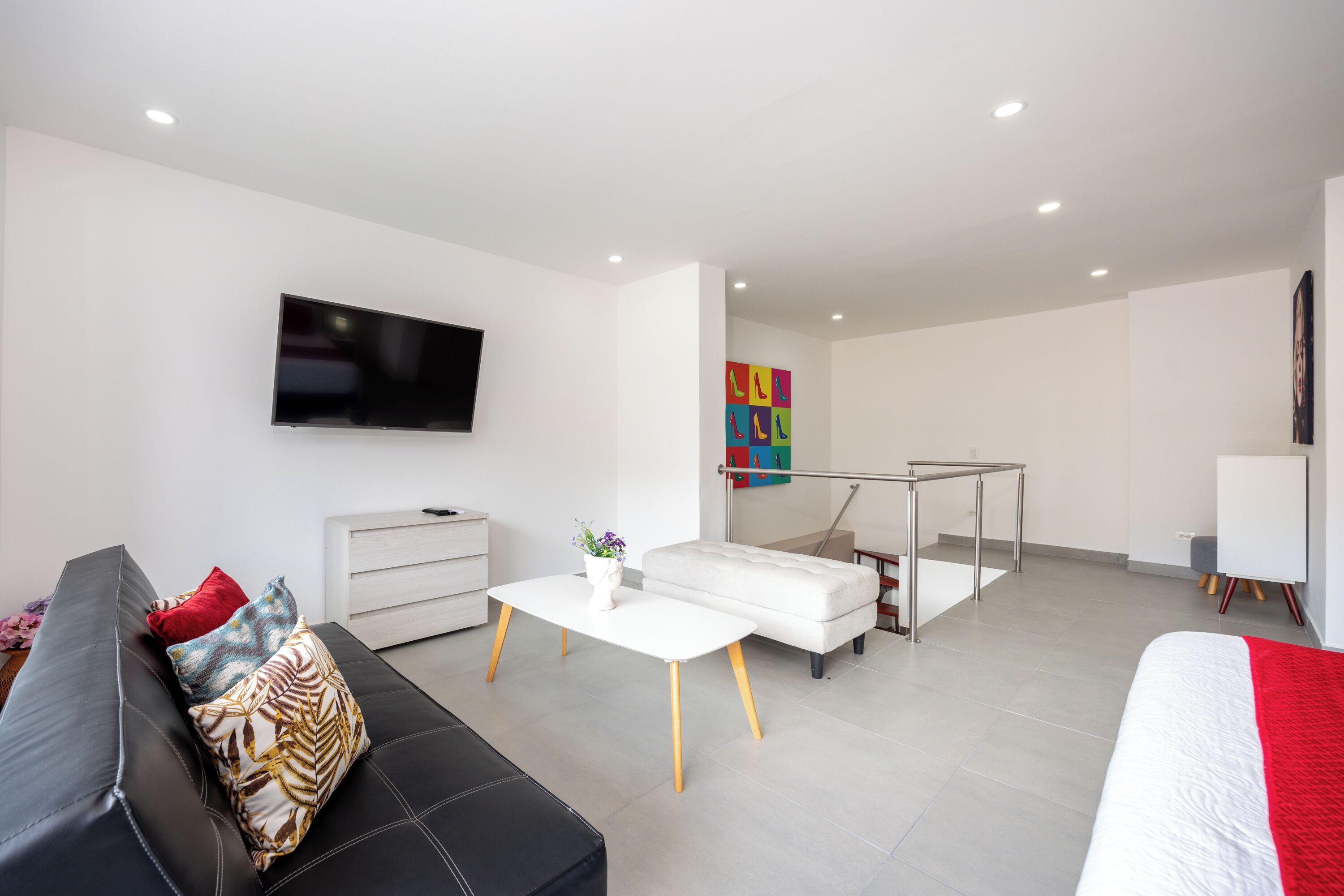 A modern living room with a black leather sofa, a white coffee table, and a white sofa. There is a flat-screen TV mounted on the wall and a small table with a vase of flowers.