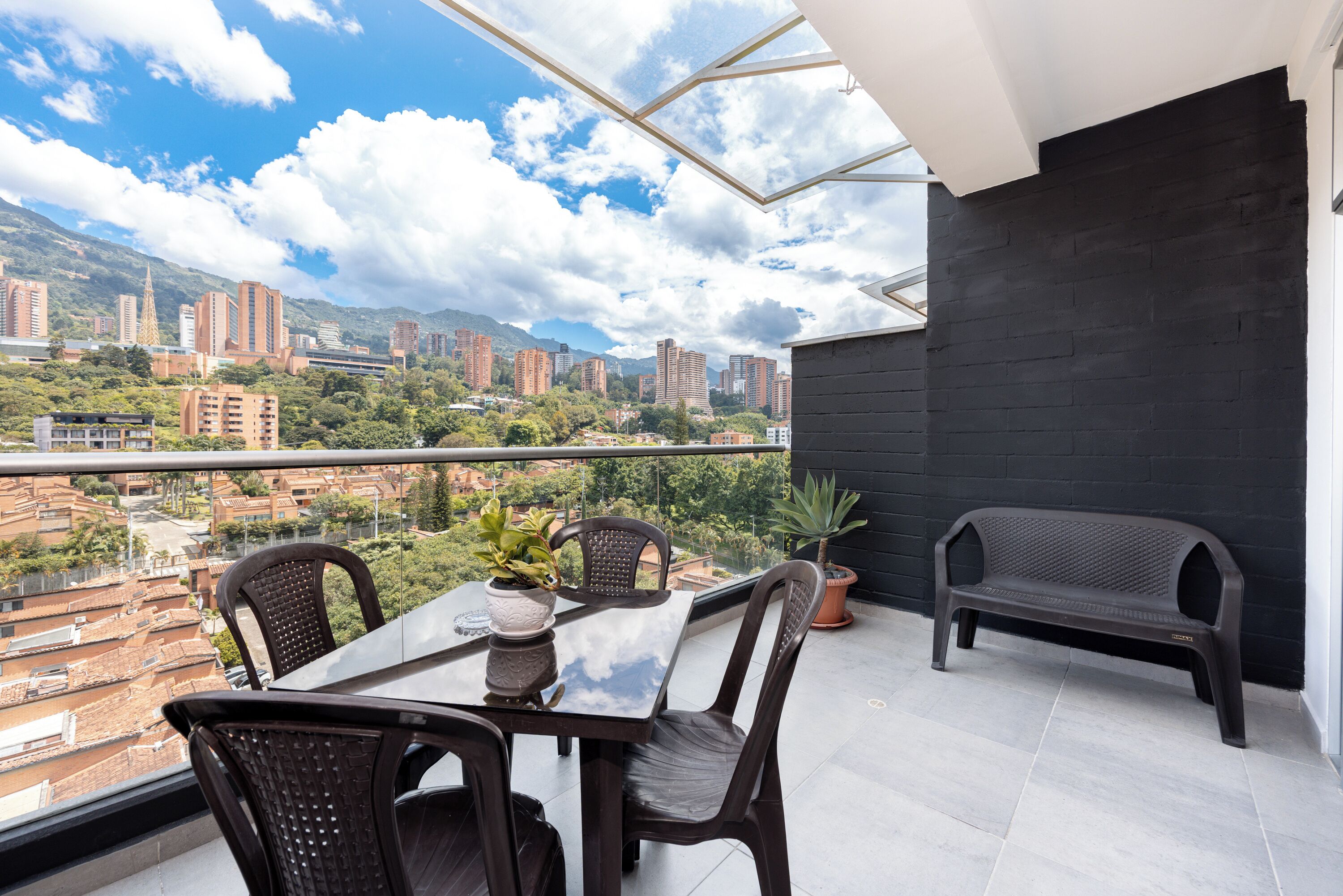 A balcony with a table and chairs, a potted plant, and a view of a cityscape with buildings and mountains.