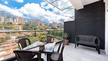 A balcony with a table and chairs, a potted plant, and a view of a cityscape with buildings and mountains.