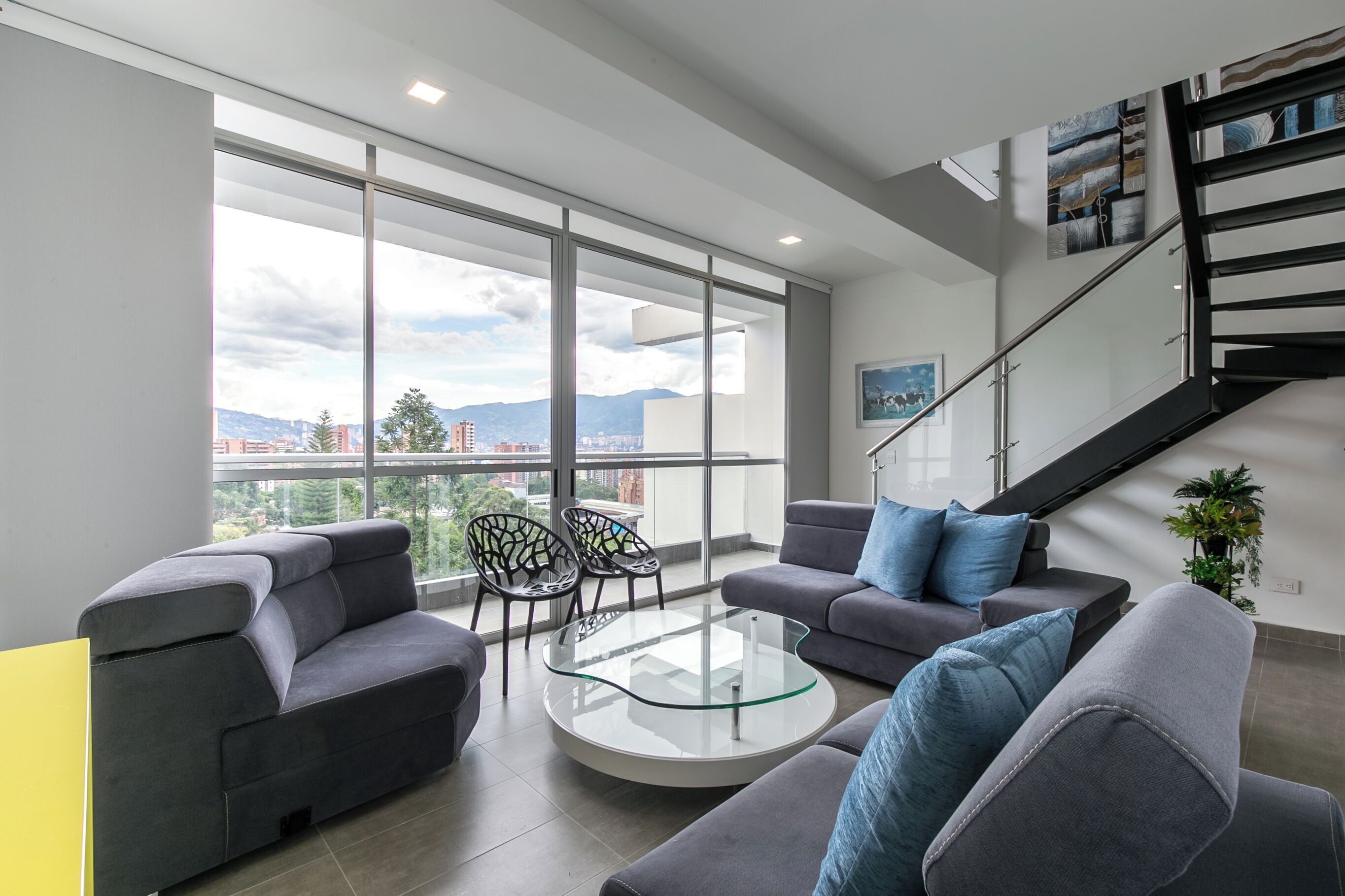 A modern living room with a glass coffee table, grey sofas, and a staircase.