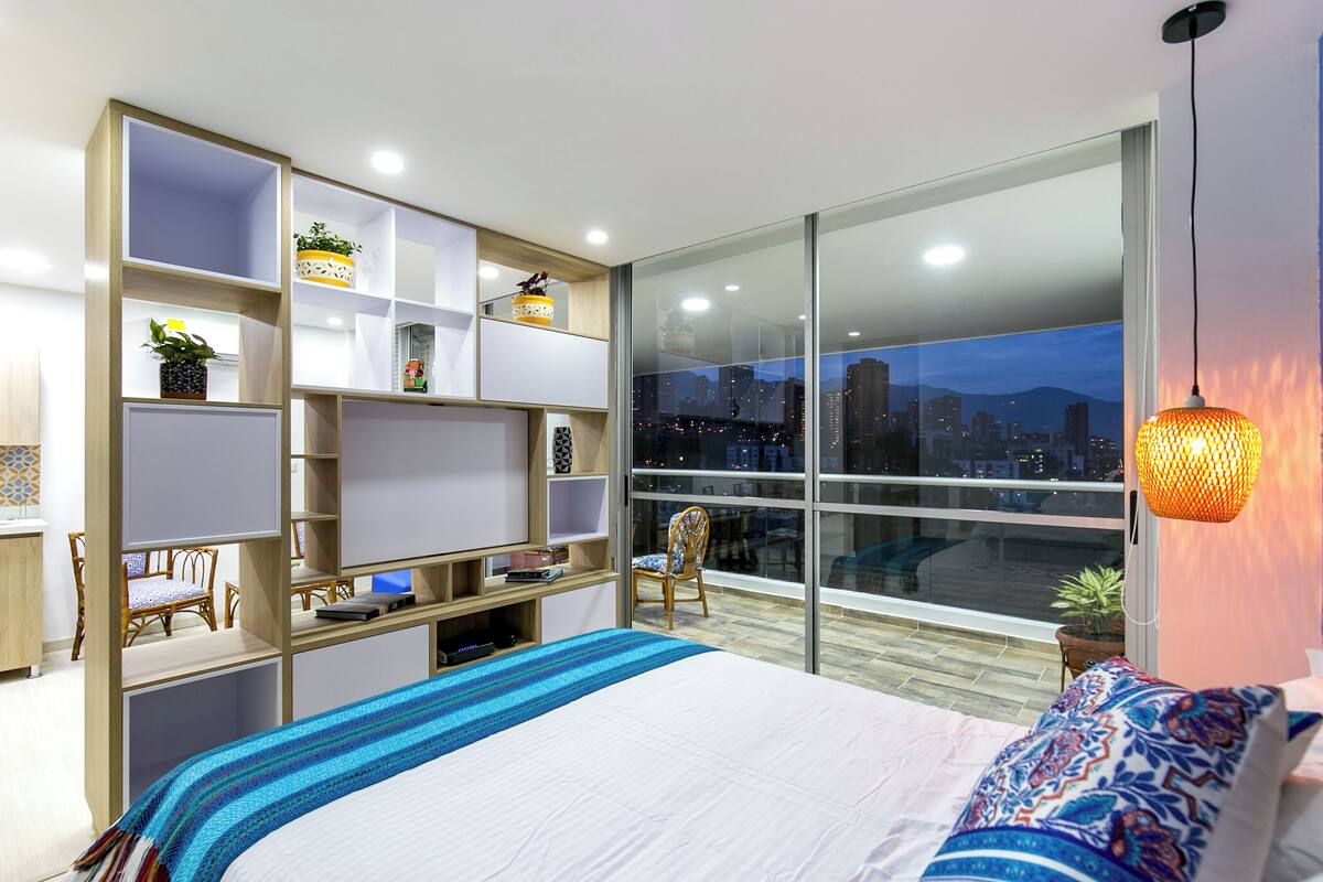 A modern bedroom with a large bed, a built-in bookshelf, a desk, and a city view through large windows.