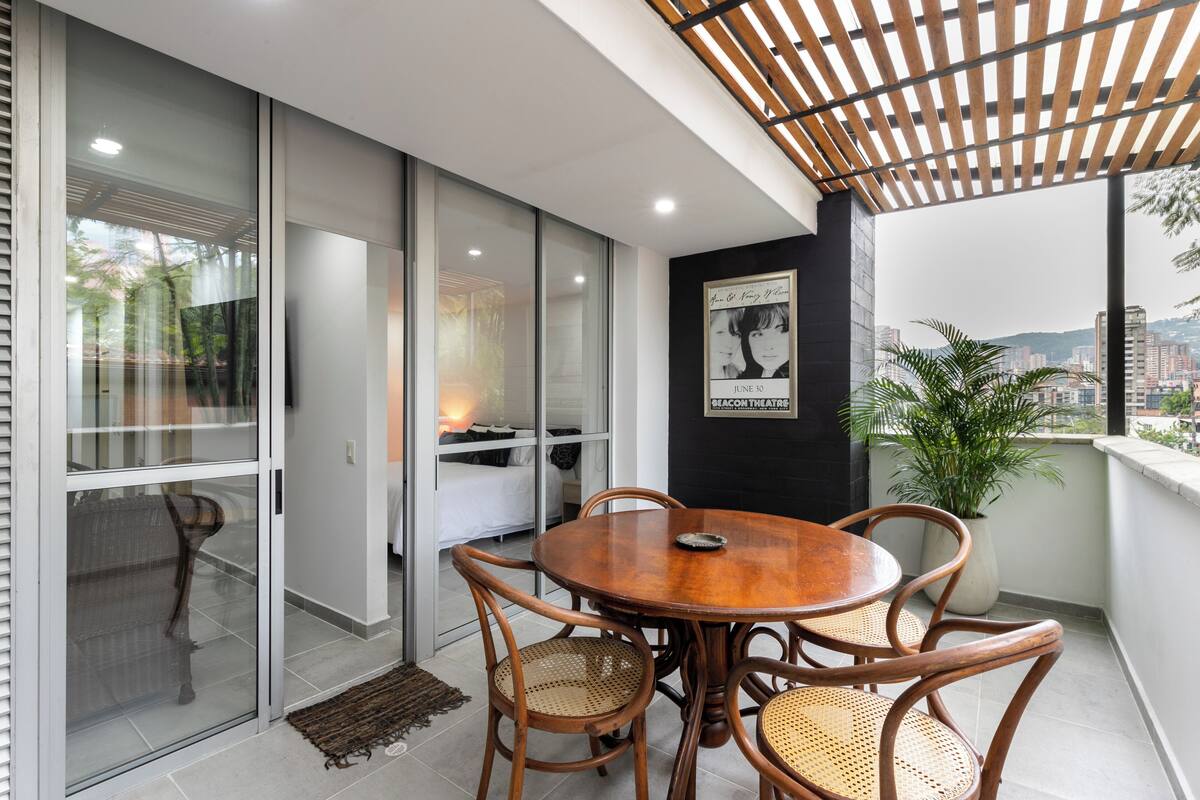 A modern balcony with a wooden table and chairs, a potted plant, and a city view.