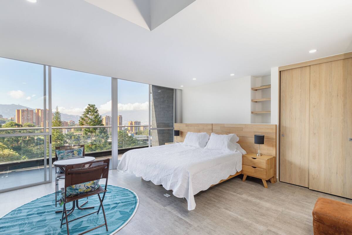 A modern bedroom with a large bed, a wooden wardrobe, a nightstand, a desk with a chair, and a view of the city through large windows.