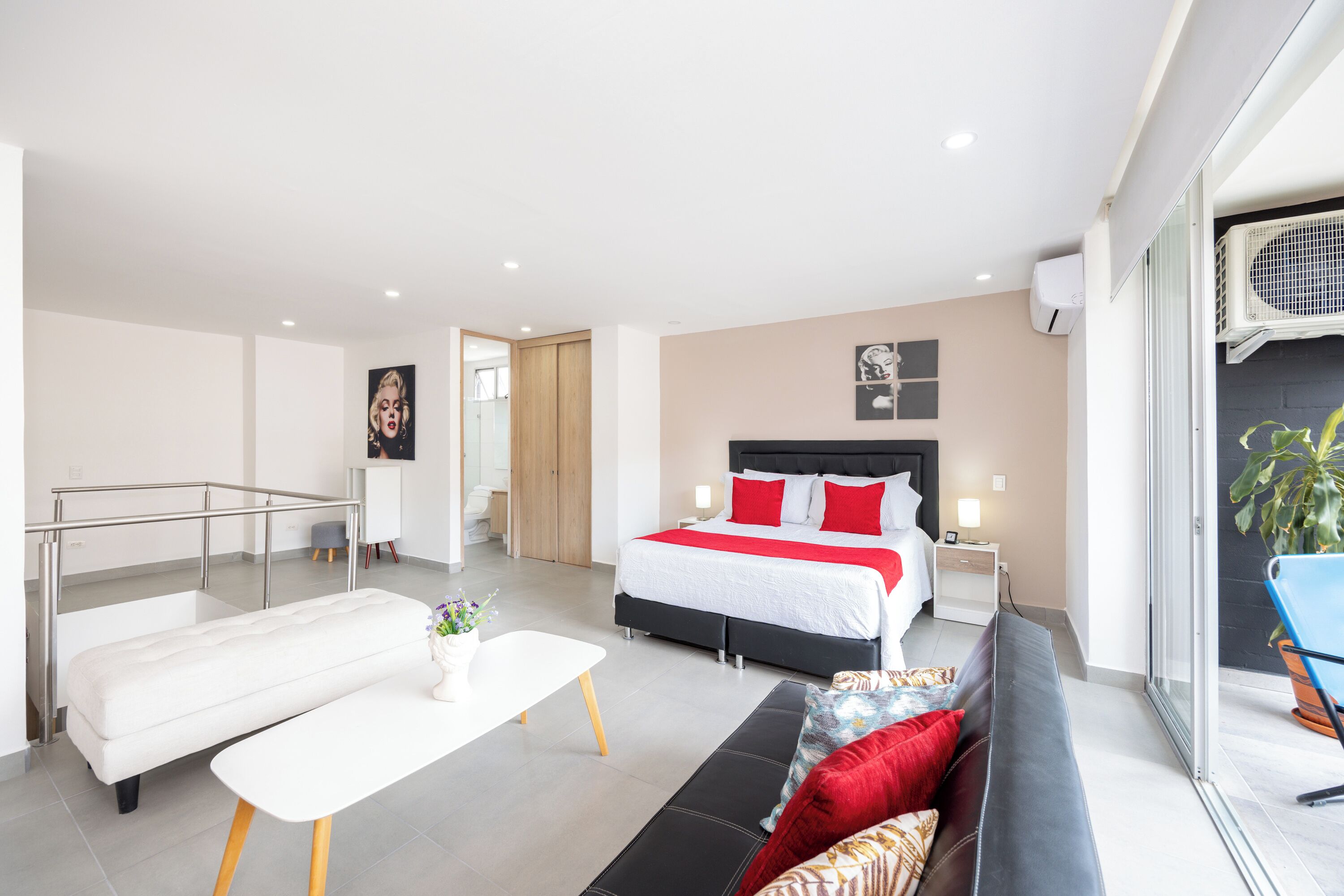 Quarto de hotel moderno com cama, sofá, mesa pequena e varanda com planta em vaso.