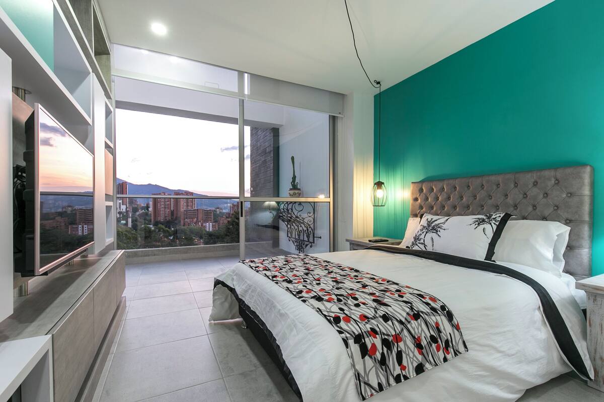A modern bedroom with a large bed, a TV, and a balcony with a city view.