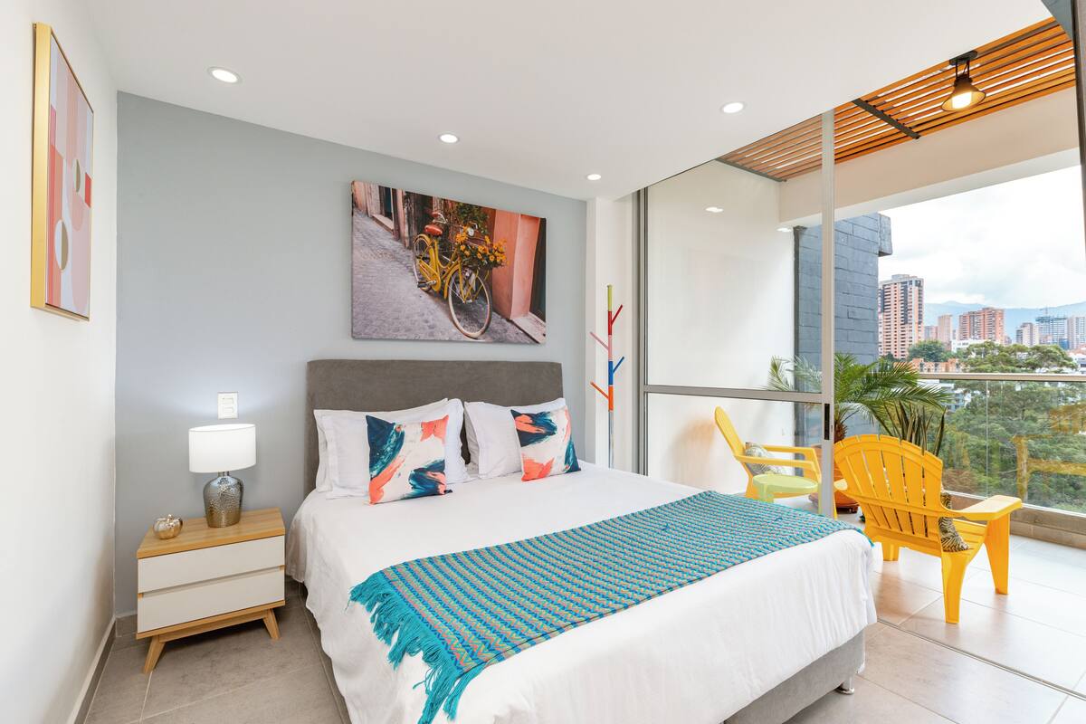A modern hotel room with a large bed, a nightstand, a chair, and a balcony with a view of the city.