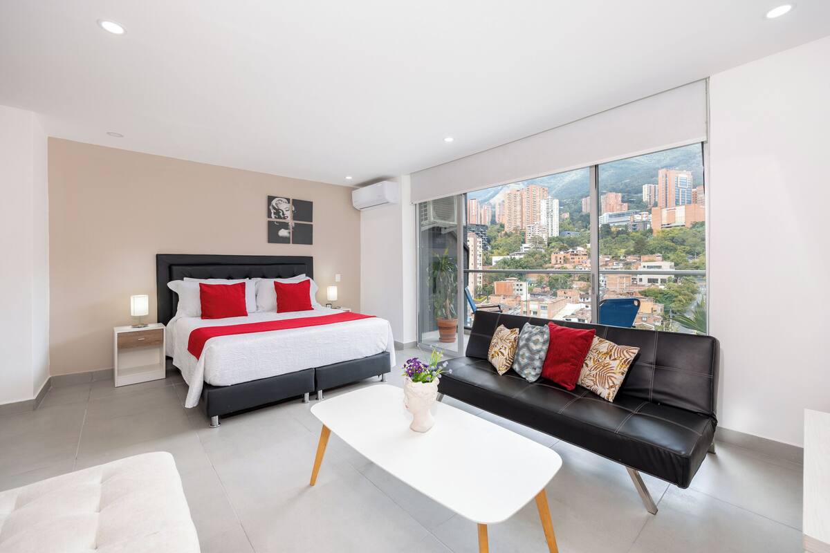 A modern hotel room with a large bed, a black sofa, a white coffee table, and a city view through large windows.