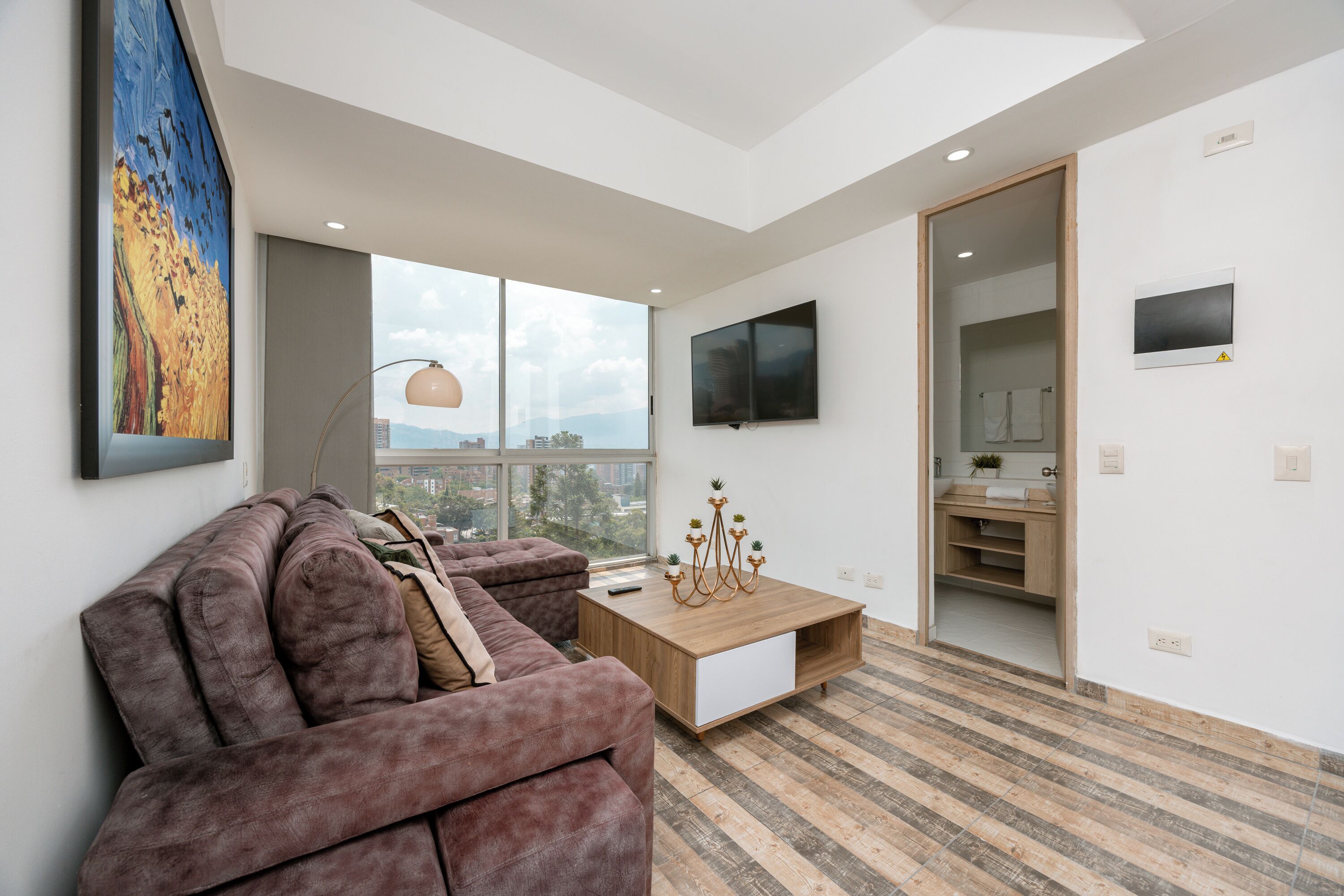 A modern living room with a large window, a sofa, a coffee table, and a TV mounted on the wall.