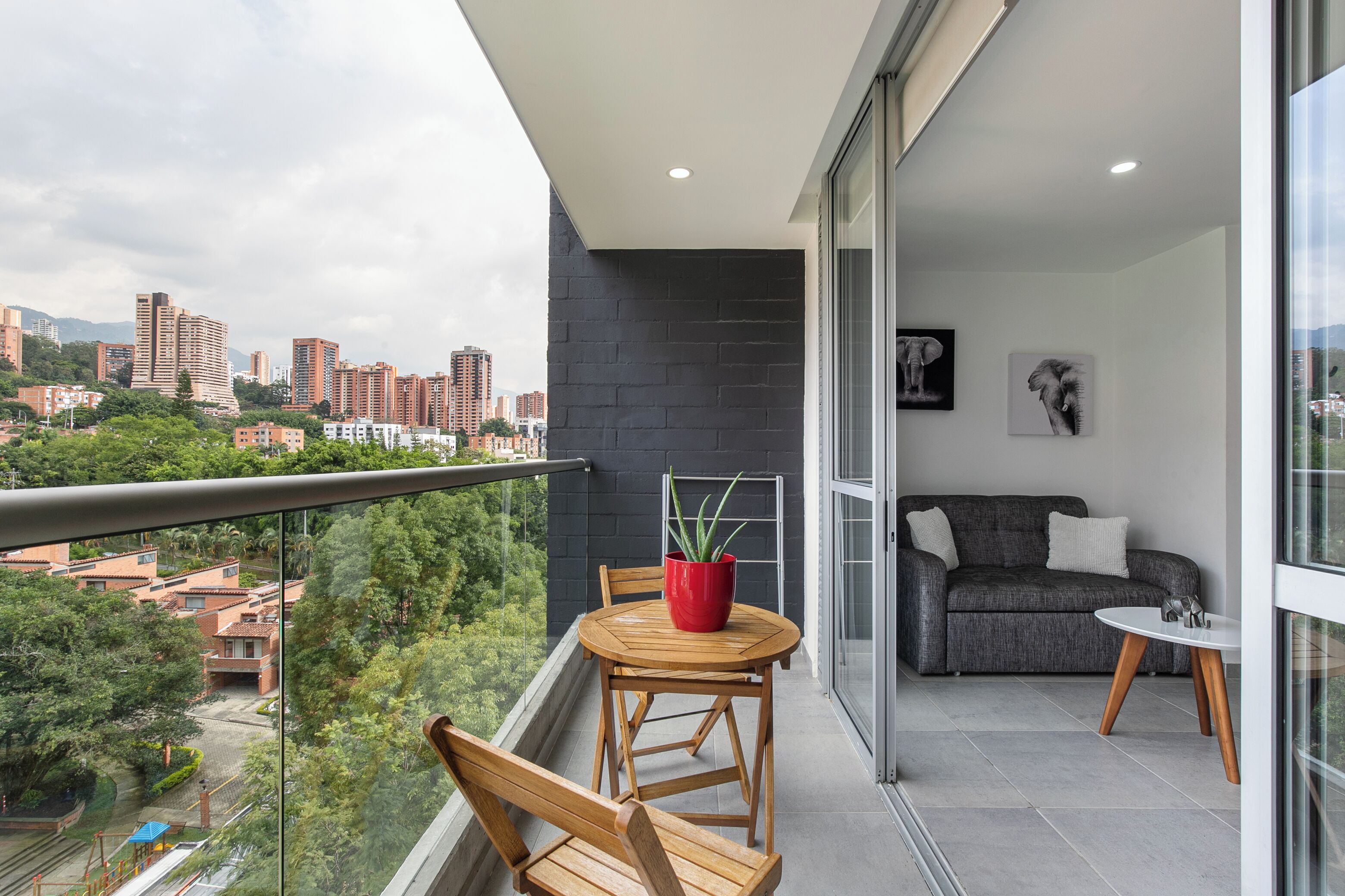 Un balcon donnant sur un paysage urbain, une plante en pot rouge, une chaise en bois, un canapé gris et une petite table blanche.