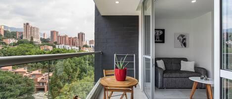 Un balcon donnant sur un paysage urbain, une plante en pot rouge, une chaise en bois, un canapé gris et une petite table blanche.