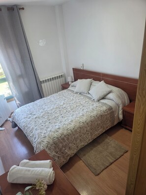 A bedroom with a bed, a nightstand, a lamp, a radiator, a window with curtains, and a wooden floor. - SNEY'S HOUSE (Sarria)