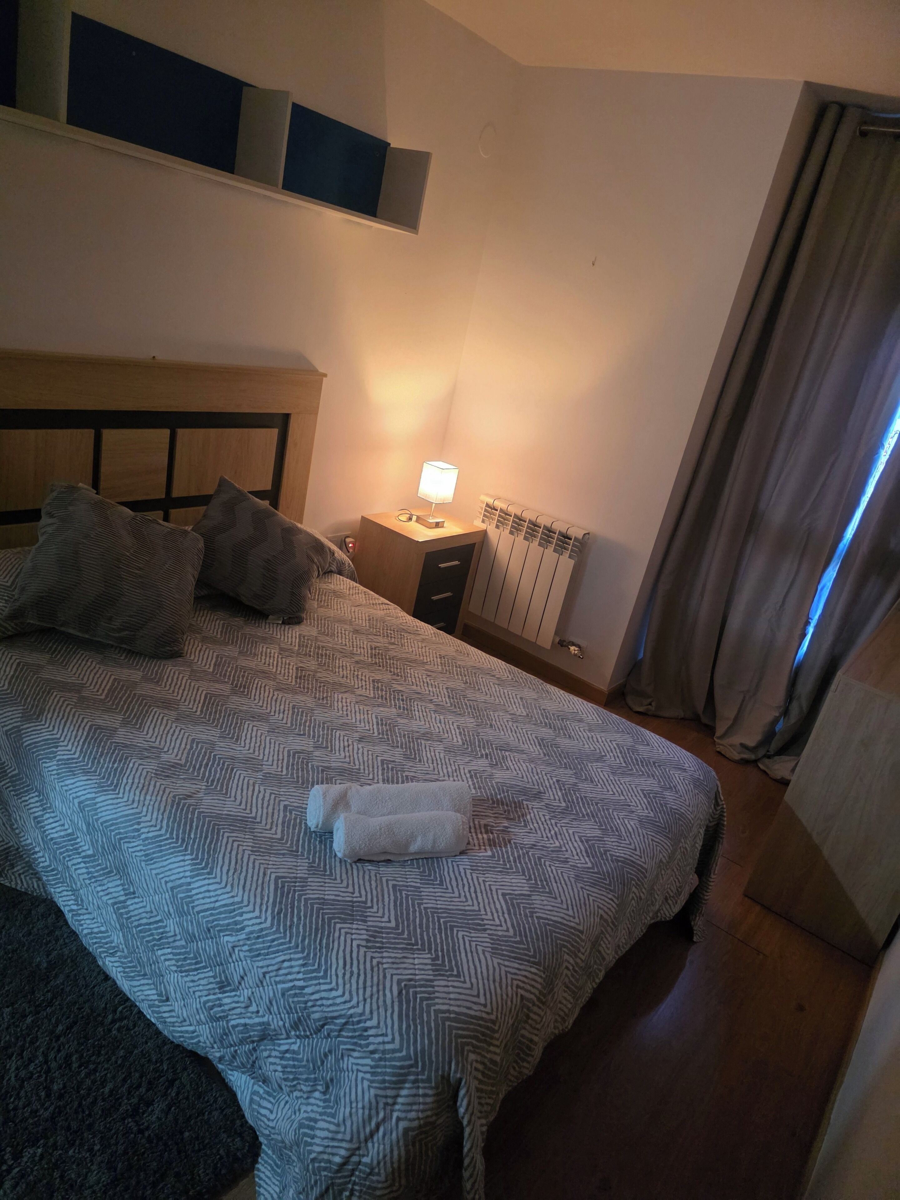 Una cama bien tendida con ropa de cama estampada, una lámpara de noche, un radiador y una ventana con cortinas.