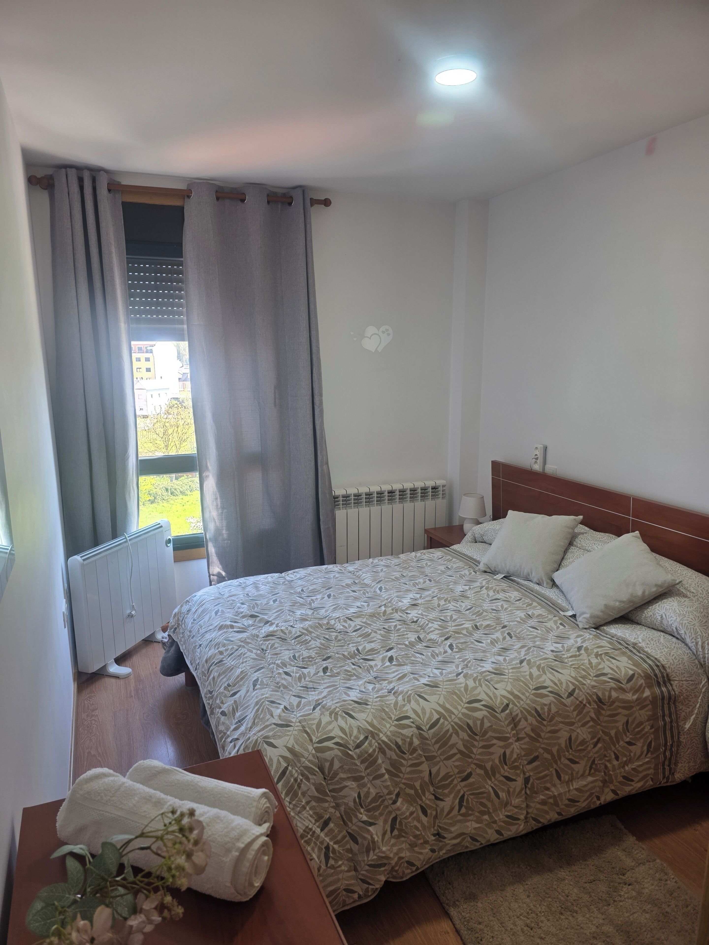 Un dormitorio con cama, un radiador, una ventana con cortinas y vistas a edificios.
