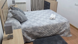 Quarto com cama, mesas de cabeceira, cômoda, radiador e janela com cortinas.