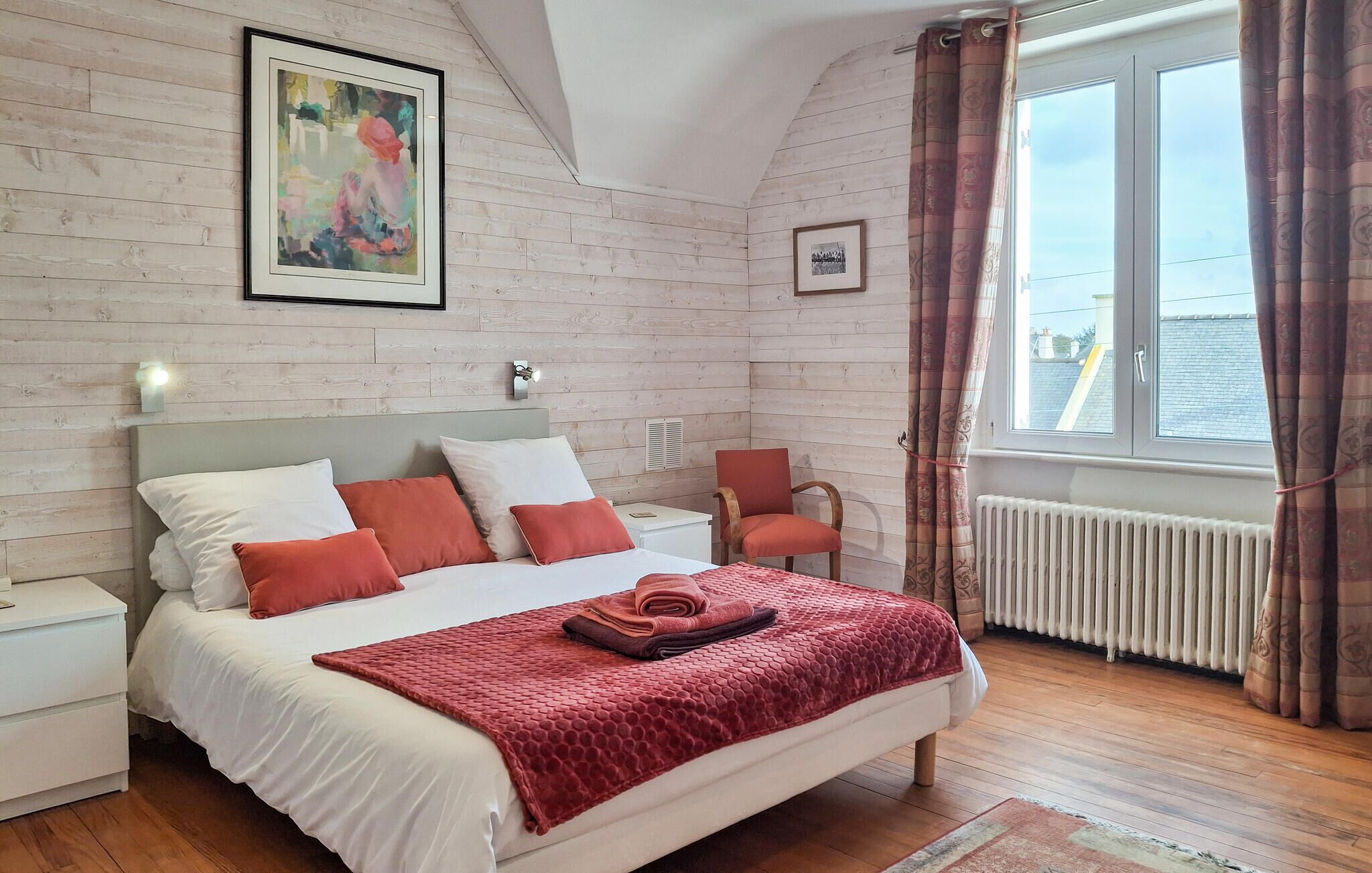 A bedroom with a bed, a nightstand, a chair, a window with curtains, and a radiator.