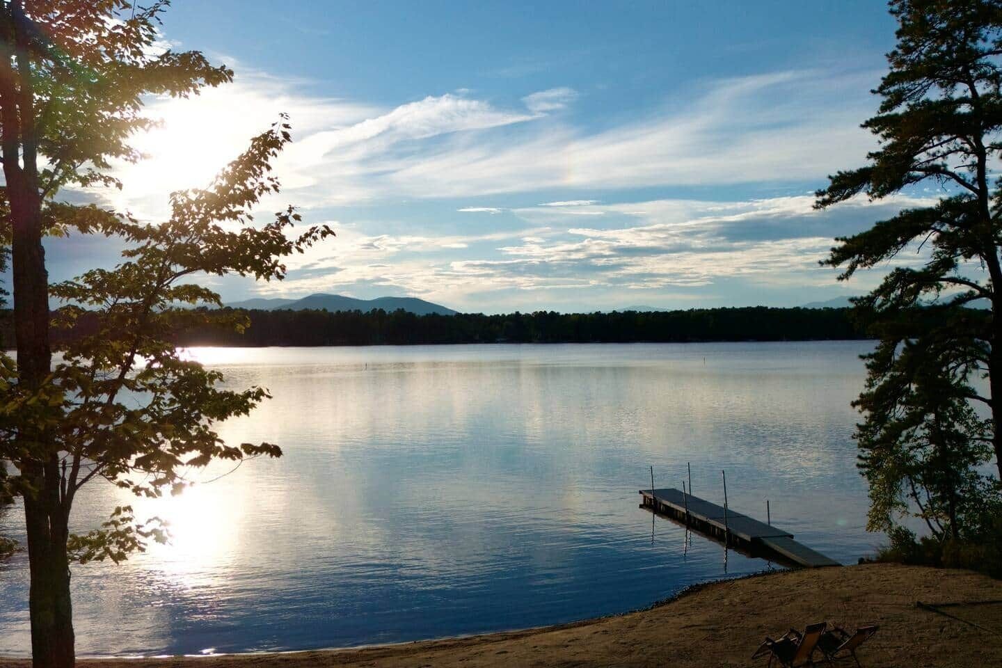 Ein ruhiger See mit einem Steg, der ins Wasser ragt, umgeben von Bäumen und Bergen im Hintergrund.