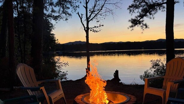 Ein Lagerfeuer mit zwei Adirondack-Stühlen vor einem ruhigen See bei Sonnenuntergang.