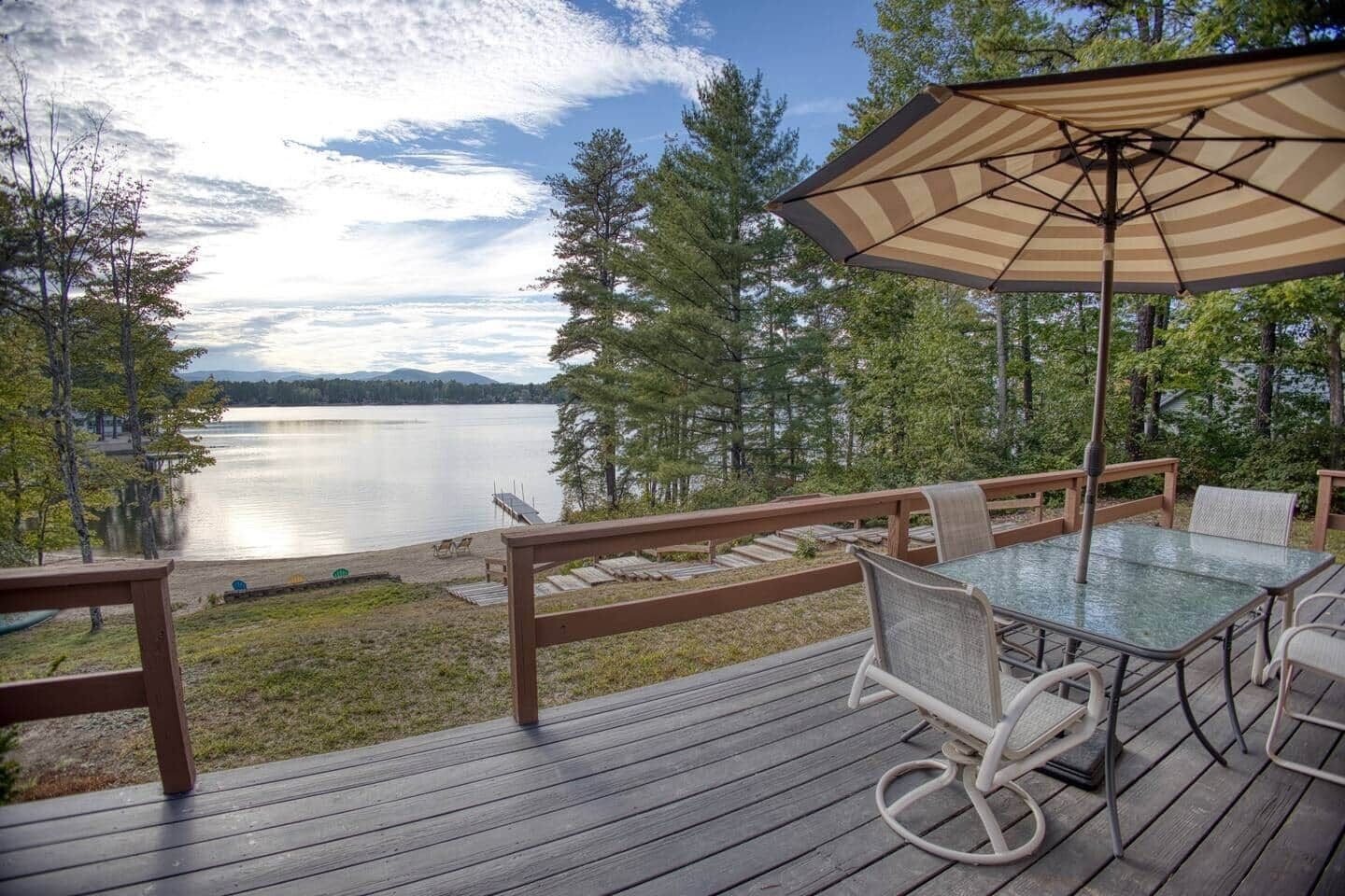 A wooden deck with a glass table and chairs, overlooking a lake with a dock and trees.