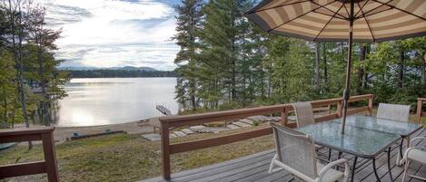 A wooden deck with a glass table and chairs, overlooking a lake with a dock and trees.