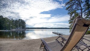 Ein Strand mit einer Holzterrasse und zwei Liegestühlen, die auf einen ruhigen See ausgerichtet sind, umgeben von Bäumen und einem Steg.