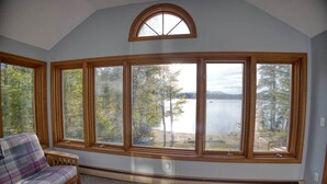 Ein Zimmer mit einem großen Erkerfenster mit Blick auf einen See und einen Wald, ein kariertes Sofa und eine hölzerne Bank.