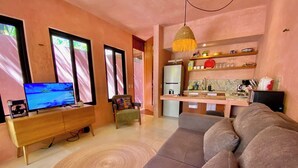 Een moderne woonkamer met een bank, televisie en een kleine fauteuil. Er is een keuken met een koelkast, gootsteen en planken.