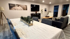 A modern living room with a long dining table, a sofa, a TV, and a staircase leading to another level.