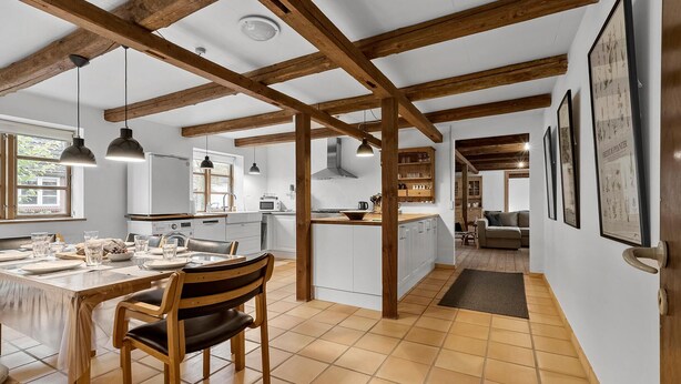 Una cocina amplia con vigas de madera, un comedor con una mesa dispuesta para cuatro personas y una sala de estar con sofá y mesa de centro.