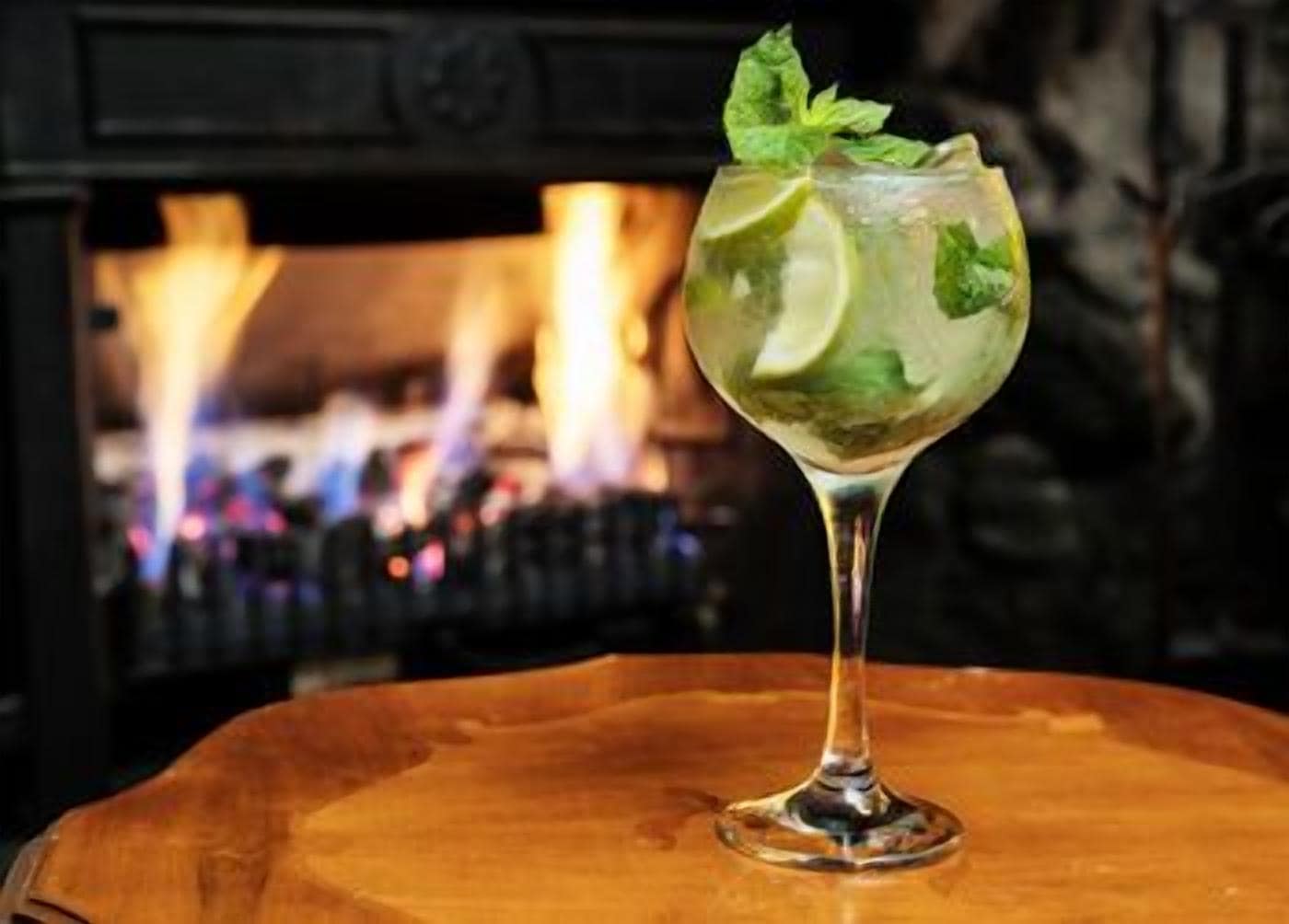 A glass with a beverage garnished with a lime and mint, placed on a wooden surface with a fireplace in the background.