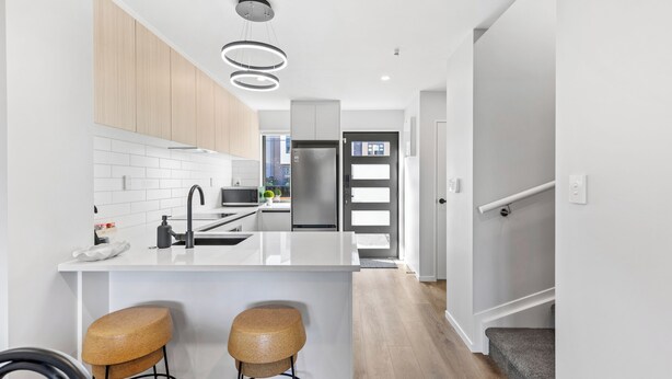 A modern kitchen with white cabinetry, a central island with stools, stainless steel appliances, and a pendant light fixture. - 4BR Townhouse for Families Zodiak Stays (Auckland)