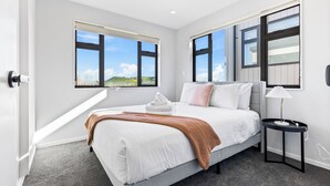 A modern bedroom with a large bed, a bedside table, and a view of the outdoors through two windows. - 4BR Townhouse for Families Zodiak Stays (Auckland)