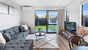 A modern living room with a grey sofa, a coffee table, a TV, and a sliding glass door leading to a balcony. - 4BR Townhouse for Families Zodiak Stays (Auckland)