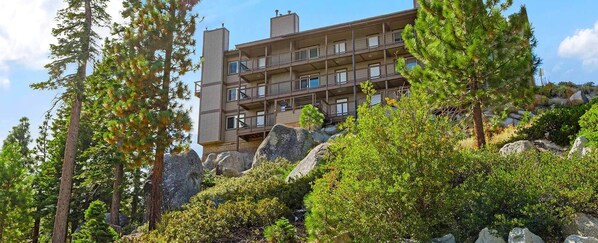 A multi-story building with balconies, surrounded by dense forest and large rocks. - Charming 2-bedroom condo with fitness room and AC in gorgeous Stateline (Stateline)