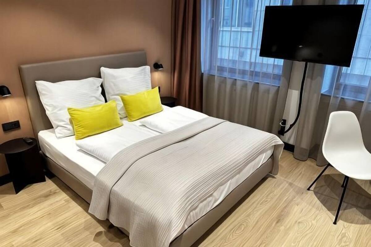 A neatly made bed with white and yellow pillows, a grey blanket, a flat-screen TV mounted on the wall, a white chair, and a small bedside table with a lamp.