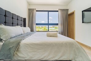 A bedroom with a large bed, a headboard, a TV mounted on the wall, and a window with curtains. - Peace on The Beach in Punta Cana (Punta Cana)