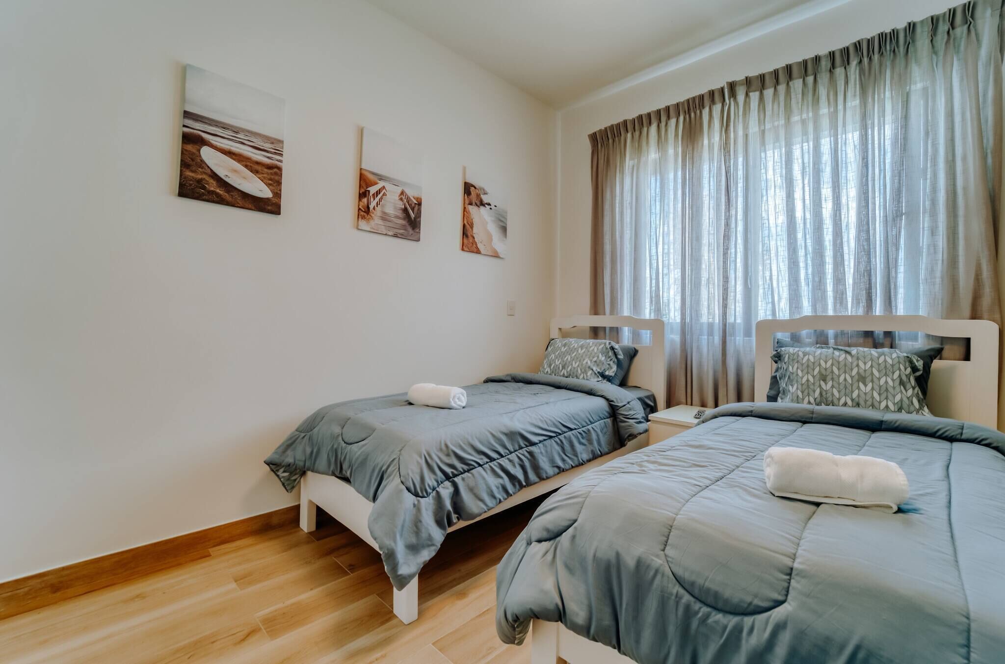 A bedroom with two beds, a large window with curtains, and three wall-mounted artworks.