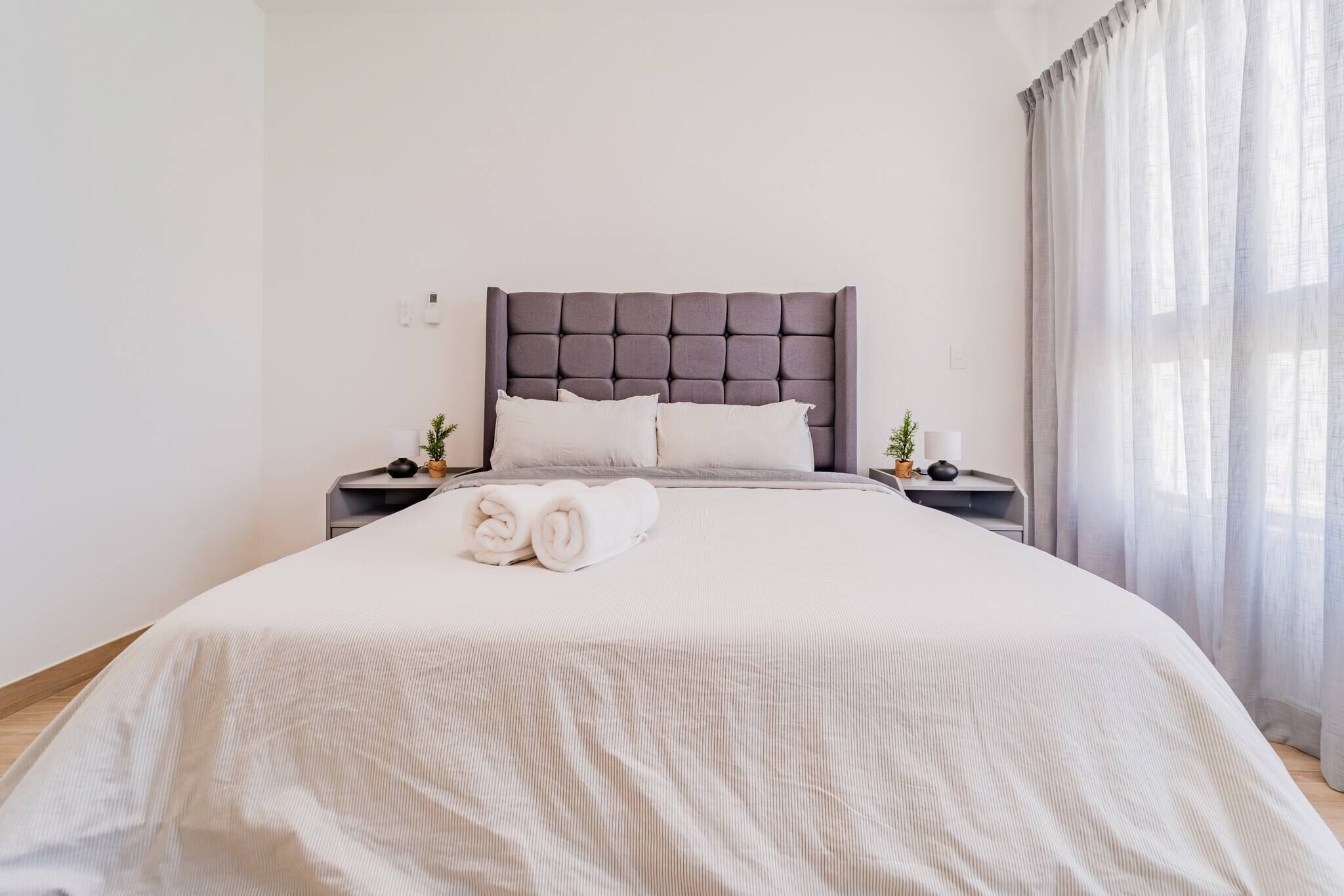 A neatly made bed with a grey headboard, white pillows, and a rolled white towel on the side.
