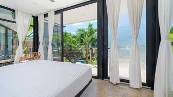 A bedroom with a large bed, a balcony with a view of the ocean, and a dining area with chairs.