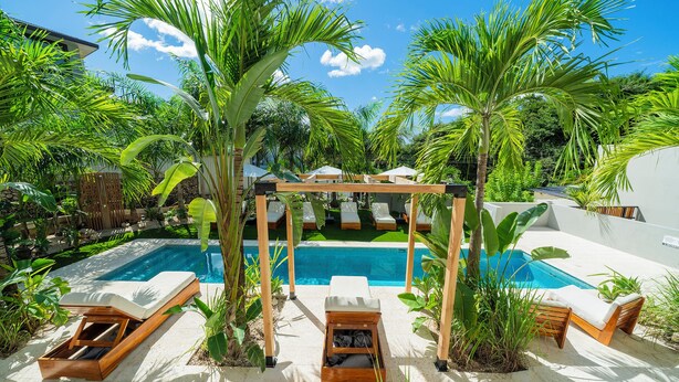 Een zwembadterras met ligbedden, een houten pergola en tropische planten.