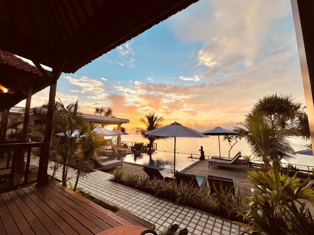 Zona de piscina en un resort con sombrillas, sillas reclinables y vista al atardecer sobre el agua.
