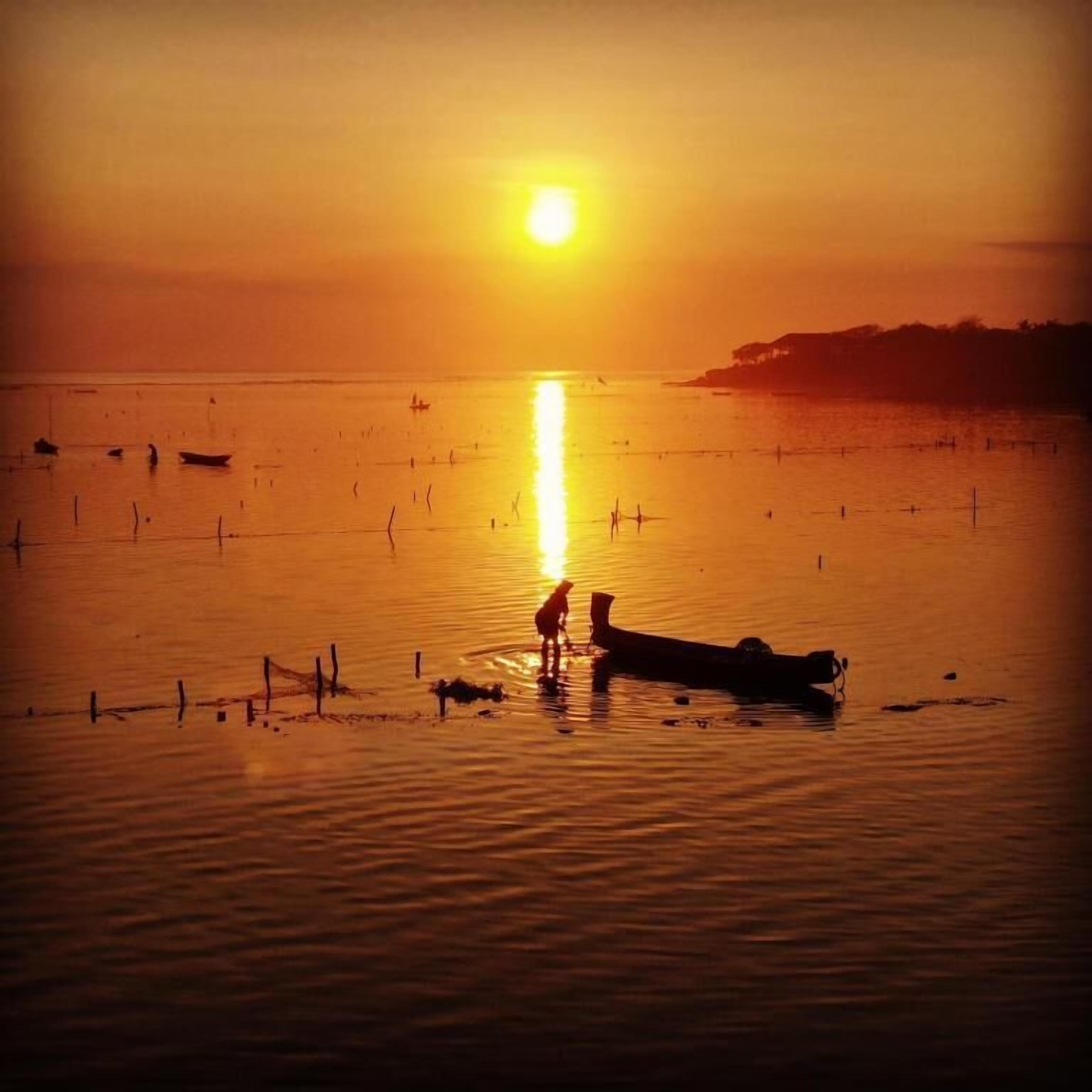 A sunset over a calm body of water with boats and a person standing on a dock.
