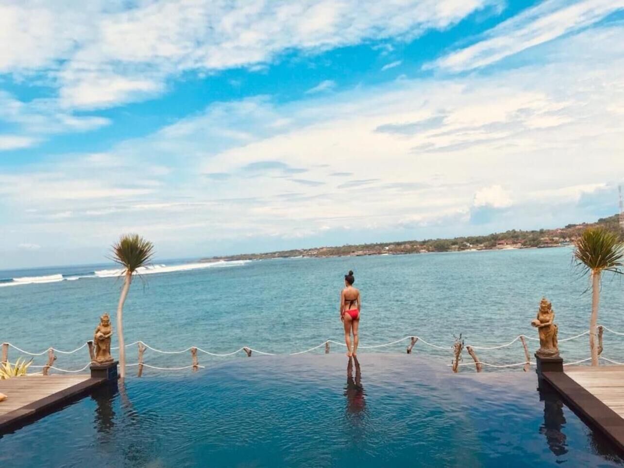 Una mujer con traje de baño rojo parada al borde de una piscina infinita con vistas al océano.