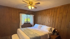Une chambre à coucher avec un lit, une table de chevet, une fenêtre avec des rideaux et un ventilateur de plafond.