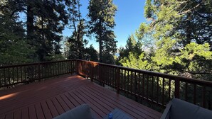 Une terrasse en bois avec une rambarde, entourée de grands arbres et surplombant un ciel bleu dégagé.