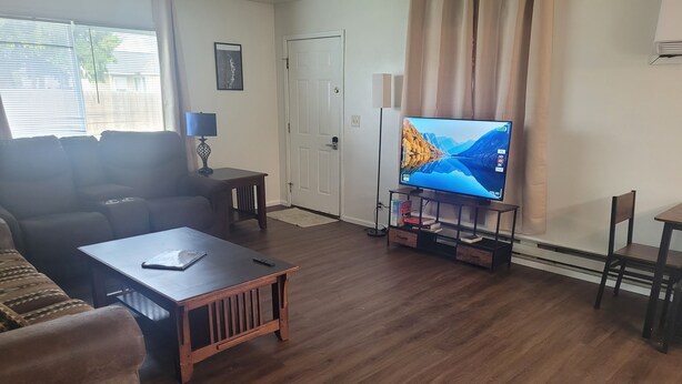 A living room with a sofa, a coffee table, a TV on a stand, a door, and a window with blinds. - Lovely family and  pet friendly townhome in charming Fruitland Idaho (Fruitland)